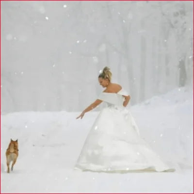 Woman in Elegant Satin Off Shoulder Bridal Gown reaching for a small dog in snow